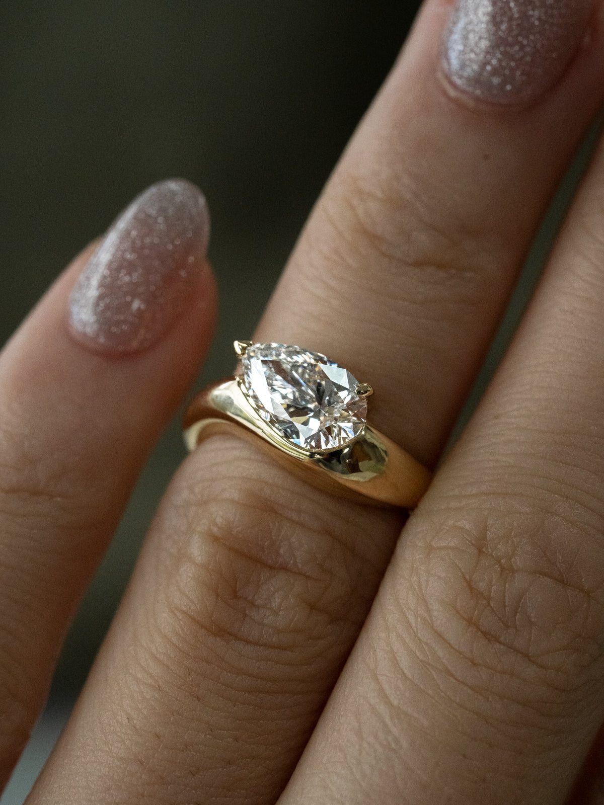 "Close-up of a pear-cut diamond engagement ring in 18k yellow gold, featuring a large brilliant diamond set in a sculptural flowing setting, shown on a person's finger with sparkly nail polish against a dark background.