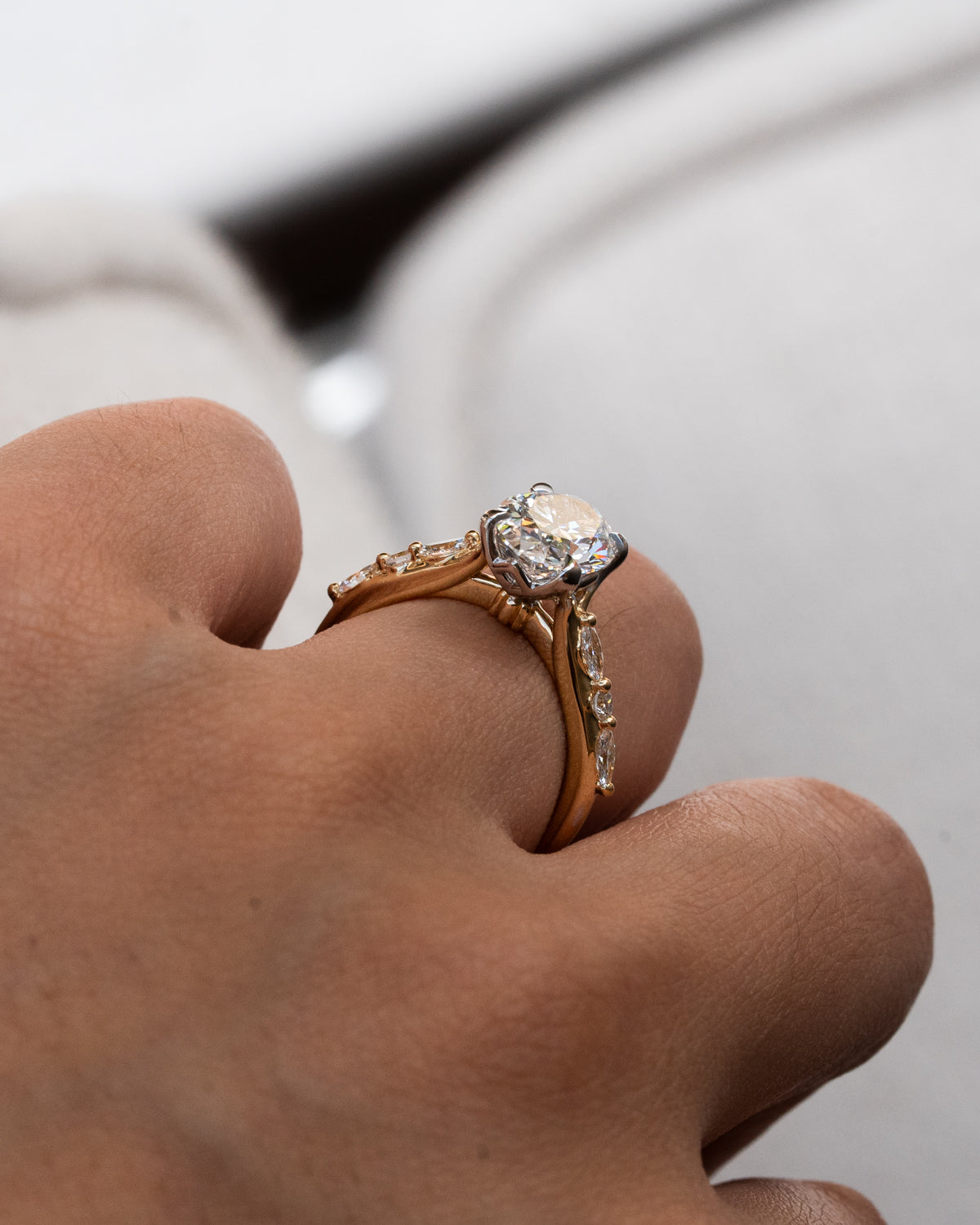 Close-up of a hand wearing a floral two toned, yellow and white gold ring with a diamond on a blurred background. Oval diamond engagement ring on hand with marquise and round diamond shoulder details
