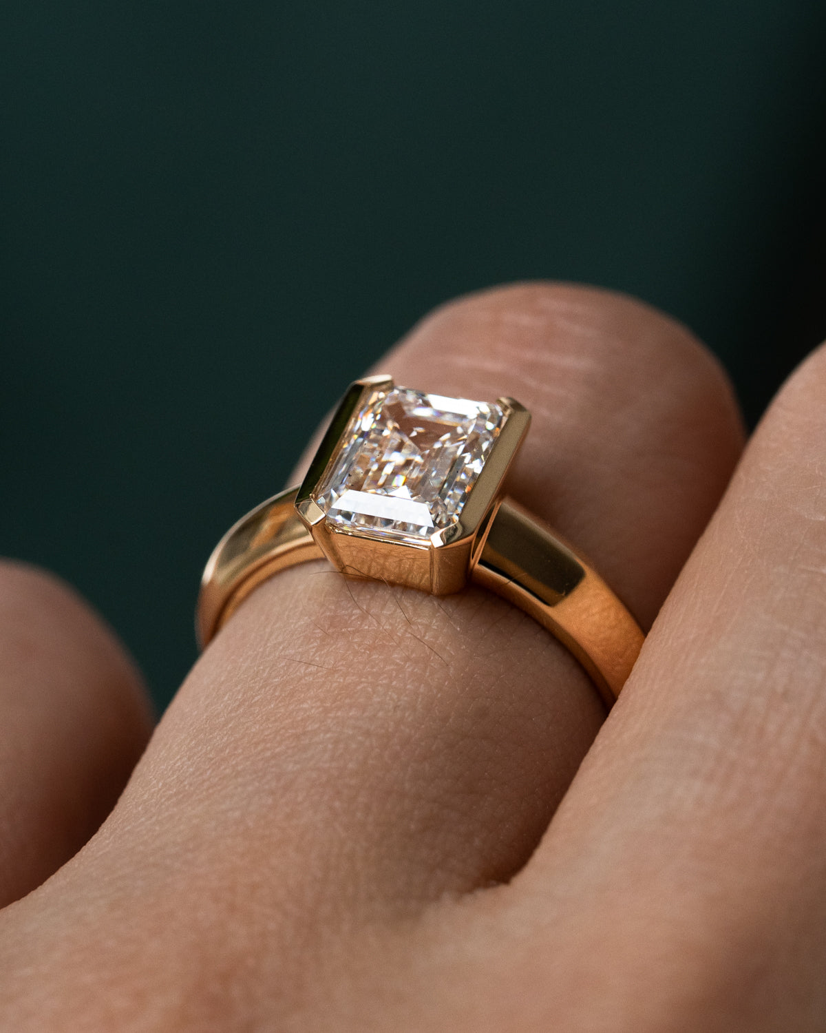 Custom yellow gold engagement ring with emerald cut diamond on hand, showing modern half bezel setting