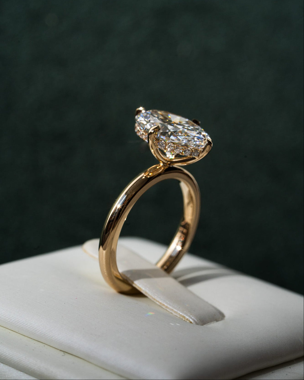A yellow gold solitaire engagement ring with a oval-cut diamond, displayed on a white jewelry stand against a dark green textured background.