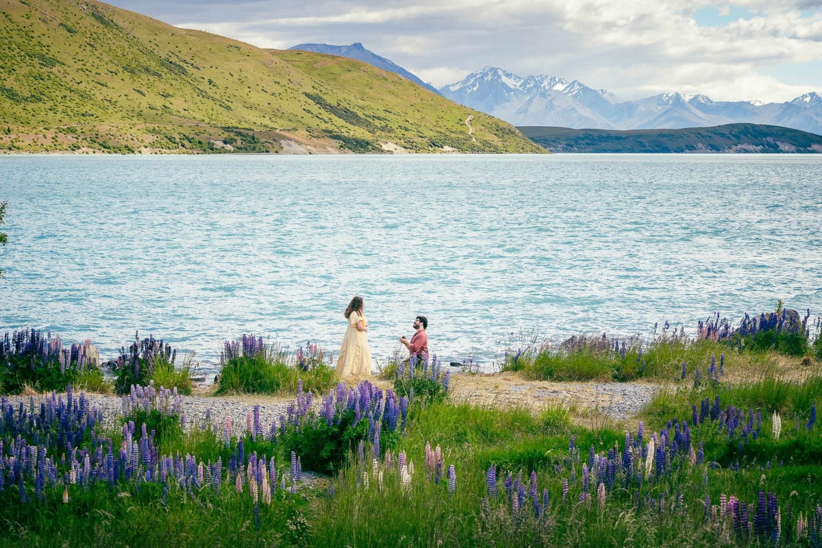 Two people standing by a lake with mountains in the background, surrounded by purple flowers.